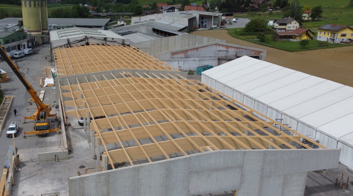 Luftaufnahme einer im Bau befindlichen Halle mit Holz-Dachkonstruktion und Betonwänden, umgeben von weiteren Hallen.