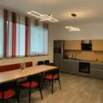 Moderner Aufenthaltsraum mit langem Holztisch, roten Stühlen und Sitzbank, großen Fenstern mit rot-weißen Lamellen und modernen Deckenleuchten.