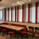 Moderner Aufenthaltsraum mit langem Holztisch, roten Stühlen und Sitzbank, großen Fenstern mit rot-weißen Lamellen und modernen Deckenleuchten.
