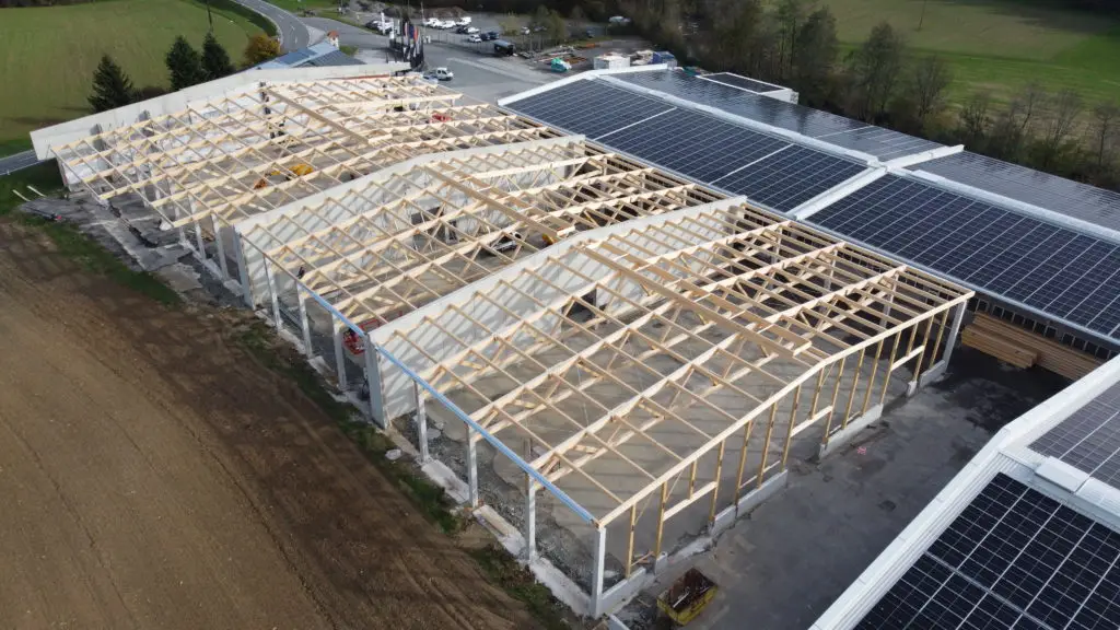 Luftaufnahme einer im Bau befindlichen Halle mit Holz-Dachkonstruktion und Betonstützen, umgeben von Feldern und Wald.