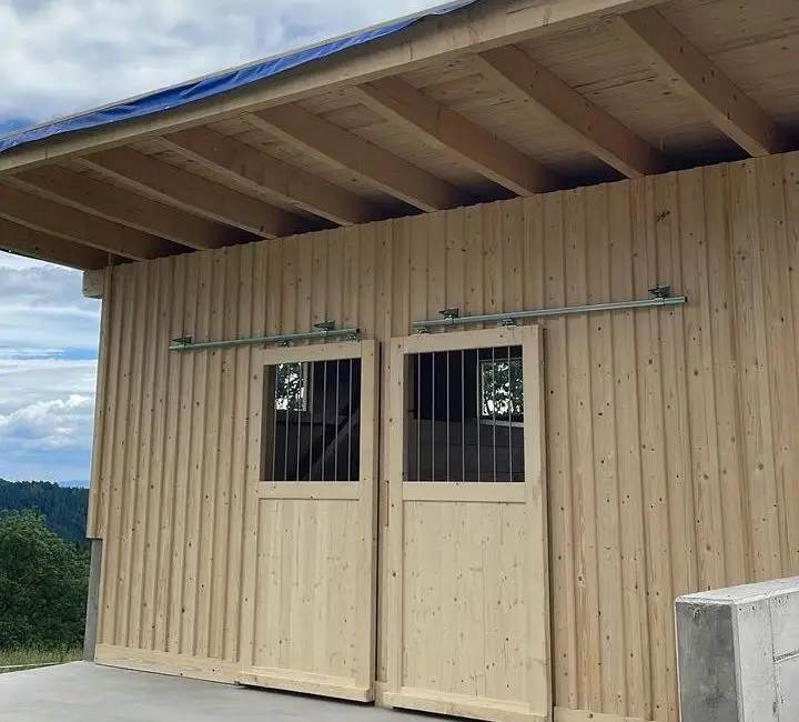 Holzgebäude mit vertikaler Holzverschalung, zwei Schiebetüren mit Metallgittern und großem Dachüberstand auf Betonboden in ländlicher Umgebung.