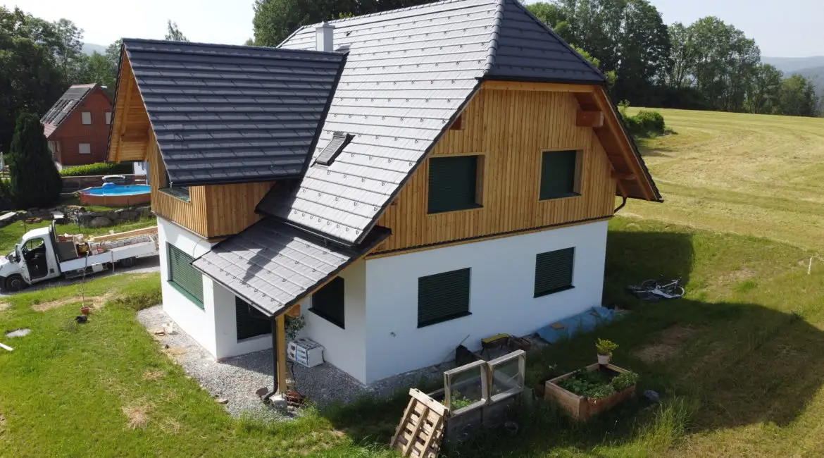 Ein modernes Einfamilienhaus auf einem leicht hügeligen Grundstück, mit weißem Putz und Holzverkleidung, dunklem Schrägdach und kleinem Balkon.