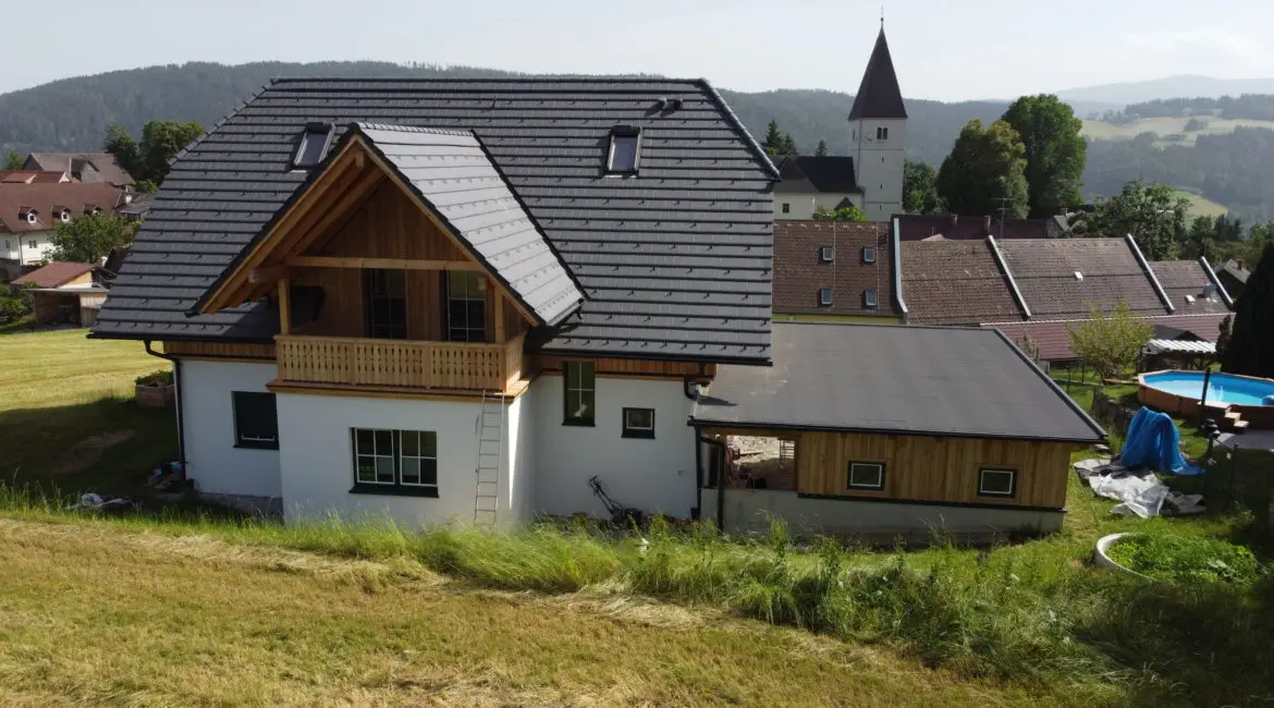 Ein modernes Einfamilienhaus auf einem leicht hügeligen Grundstück, mit weißem Putz und Holzverkleidung, dunklem Schrägdach und kleinem Balkon. Rechts daneben befindet sich ein flacherer Anbau. Im Hintergrund sind weitere Häuser, ein Kirchturm und bewaldete Hügel zu sehen. Auf der rechten Seite ist ein runder Swimmingpool mit blauer Abdeckung.