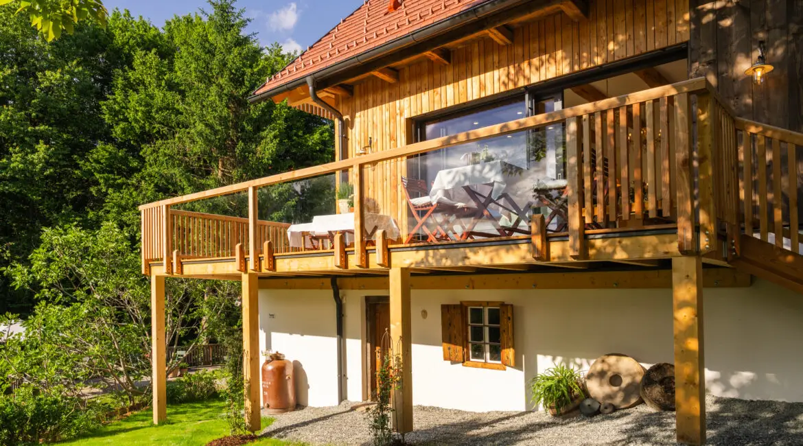 Holzhaus mit rotem Ziegeldach und Terrasse, gedeckte Tische vor großer Glasfront, eingebettet in grünen Garten.