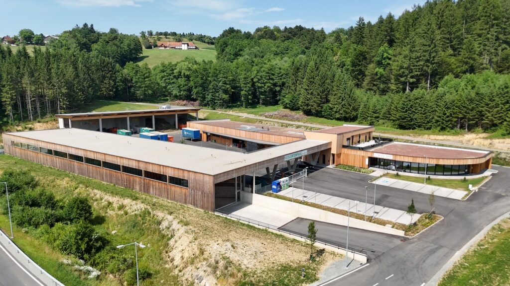 Luftaufnahme eines modernen Recyclingzentrums mit Holzfassaden, umgeben von grünen Hügeln und Waldlandschaft.