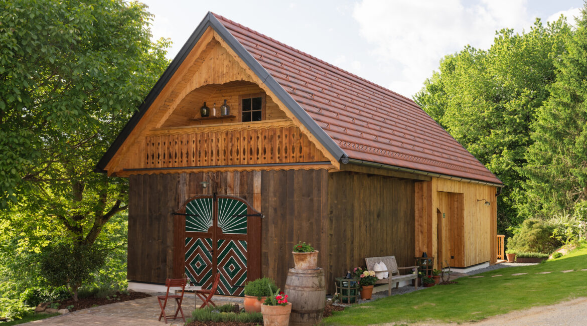 Rustikales Holzhaus mit Satteldach, grünen Scheunentoren und Blumen vor der Tür, umgeben von Bäumen und Wiese.