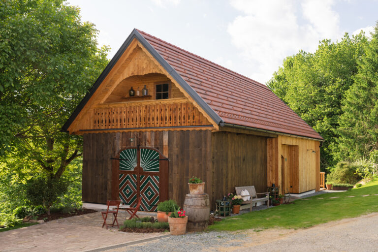 Rustikales Holzhaus mit Satteldach, grünen Scheunentoren und Blumen vor der Tür, umgeben von Bäumen und Wiese.