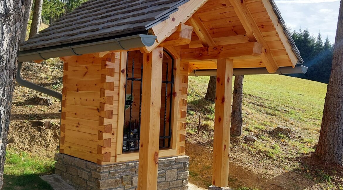 Kleine Holzkapelle mit Vorbau im Wald, auf Steinsockel gebaut, umgeben von Bäumen und Wiese bei sonnigem Wetter.