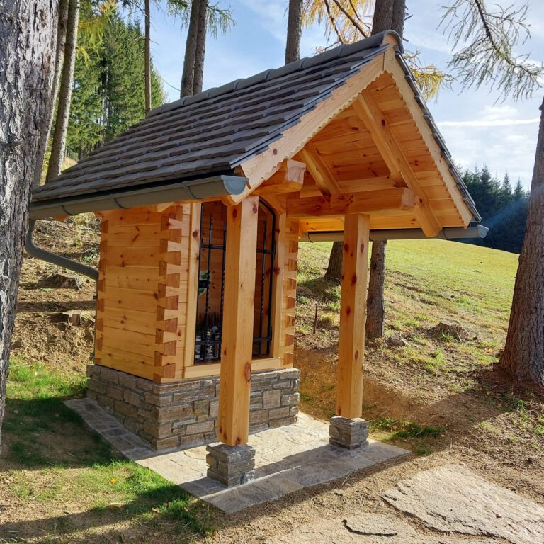 Kleine Holzkapelle mit Vorbau im Wald, auf Steinsockel gebaut, umgeben von Bäumen und Wiese bei sonnigem Wetter.