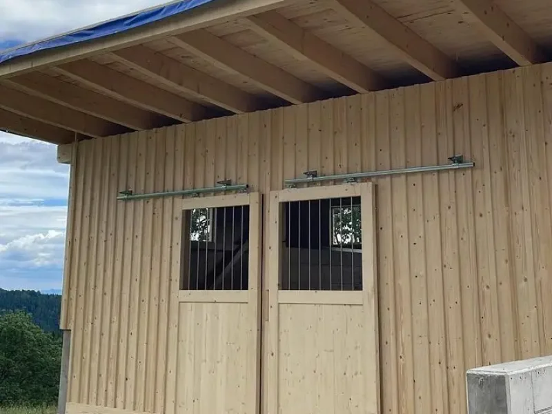 Holzgebäude mit vertikaler Holzverschalung, zwei Schiebetüren mit Metallgittern und großem Dachüberstand auf Betonboden in ländlicher Umgebung.