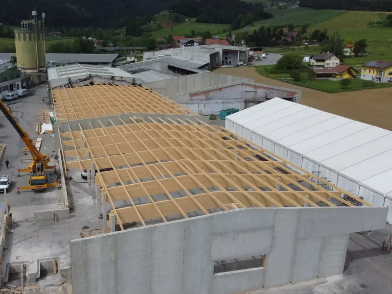 Luftaufnahme einer im Bau befindlichen Halle mit Holz-Dachkonstruktion und Betonwänden, umgeben von weiteren Hallen.