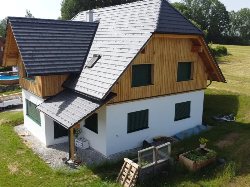 Ein modernes Einfamilienhaus auf einem leicht hügeligen Grundstück, mit weißem Putz und Holzverkleidung, dunklem Schrägdach und kleinem Balkon.