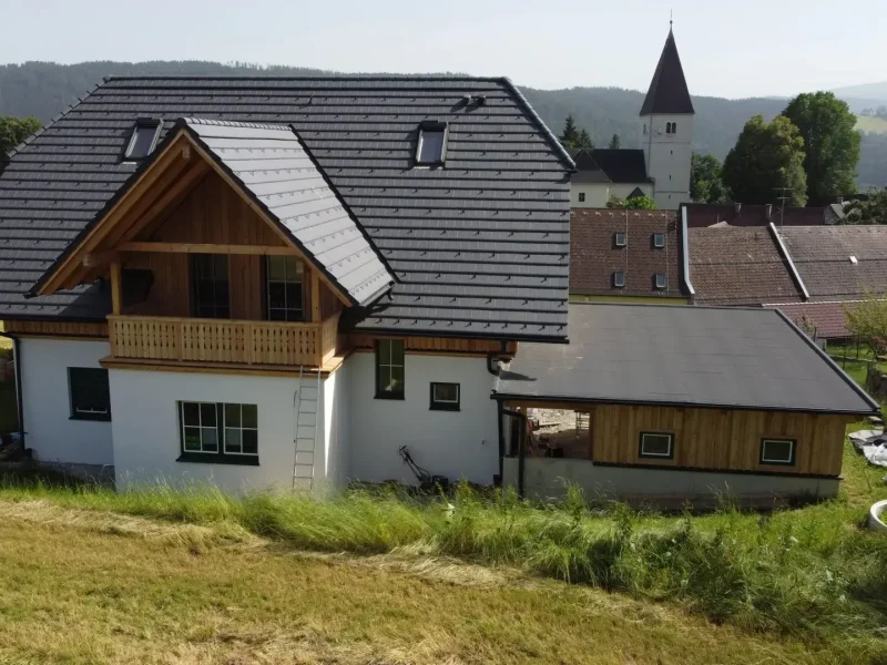 Ein modernes Einfamilienhaus auf einem leicht hügeligen Grundstück, mit weißem Putz und Holzverkleidung, dunklem Schrägdach und kleinem Balkon. Rechts daneben befindet sich ein flacherer Anbau. Im Hintergrund sind weitere Häuser, ein Kirchturm und bewaldete Hügel zu sehen. Auf der rechten Seite ist ein runder Swimmingpool mit blauer Abdeckung.