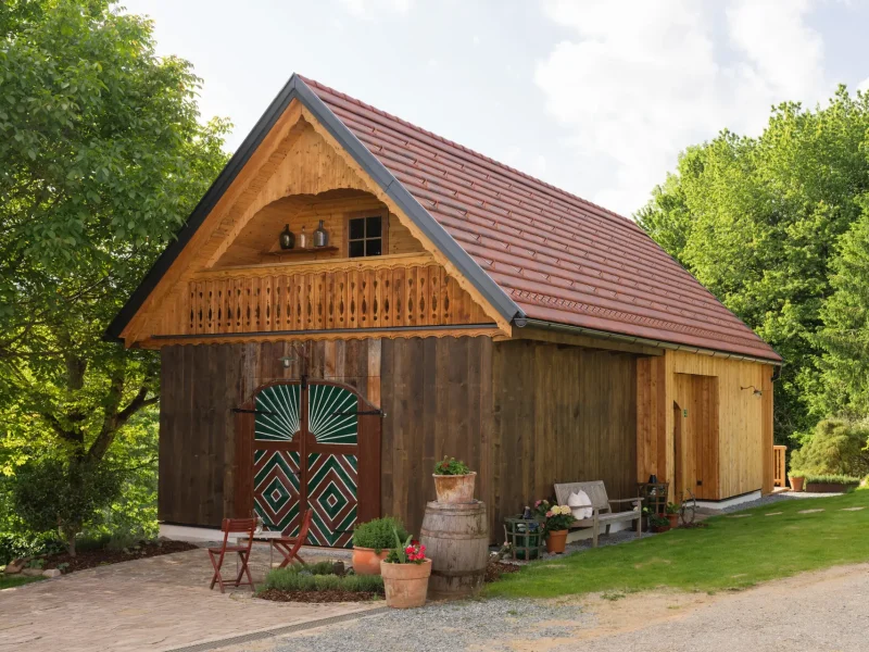 Rustikales Holzhaus mit Satteldach, grünen Scheunentoren und Blumen vor der Tür, umgeben von Bäumen und Wiese.