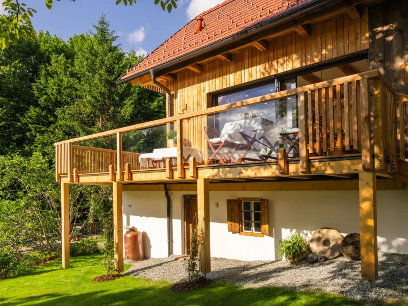 Holzhaus mit rotem Ziegeldach und Terrasse, gedeckte Tische vor großer Glasfront, eingebettet in grünen Garten.