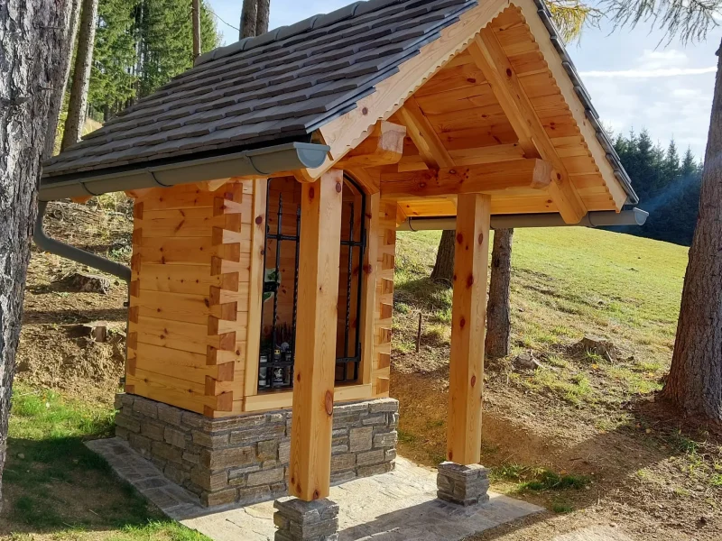 Kleine Holzkapelle mit Vorbau im Wald, auf Steinsockel gebaut, umgeben von Bäumen und Wiese bei sonnigem Wetter.